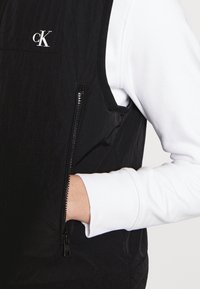 Czarna kamizelka bez rękawów o pikowanej strukturze, z zamykaną na zamek kieszenią i małym białym logo na piersi, noszona na białej koszuli z długim rękawem.