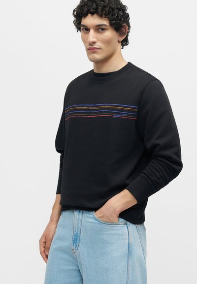 Νέος άνδρας με σγουρά μαλλιά, που φοράει μαύρο φούτερ με πολύχρωμες οριζόντιες γραμμές και ανοιχτό μπλε τζιν, με το χέρι στην τσέπη, κοιτάζοντας εμπρός.