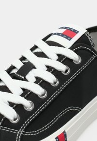 Μαύρο αθλητικό παπούτσι από καμβά με λευκά κορδόνια, ασημένια μεταλλικά τρύπες και αντίθετες λευκές ραφές. Διαθέτει λογότυπο σε κόκκινο, λευκό και μπλε χρώμα.