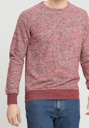 Uomo che indossa un maglione lavorato a maglia rosso e grigio melange con polsini e orlo a coste, abbinato a jeans blu, su uno sfondo semplice.