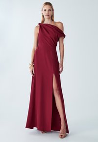 Burgundia tooni ühe õlaga kleit siledas kangas, millel on kõrge lõhik ja õlal drapeering, kombineeritud neutraalsete sandaalidega.