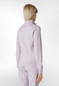 Ljuslila zip-up jacka med hög krage, ribbade muddar och en liten rosa logotyp på baksidan. Slät textur och tätt passform.