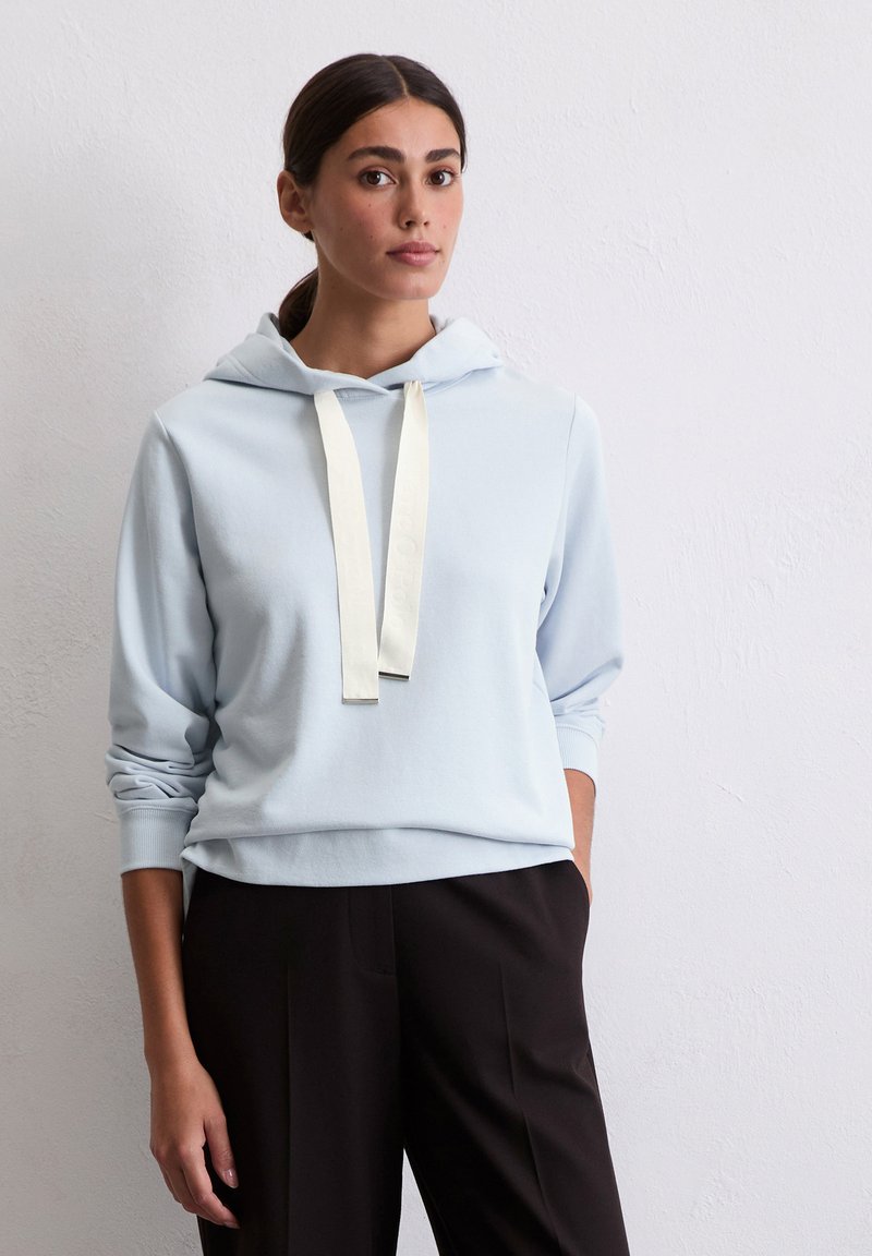 Helles blaues Hoodie mit einer frontalen Kängurutasche, ausgestattet mit einer cremefarbenen Kordel und gerippten Bündchen, kombiniert mit schwarzen Hosen.