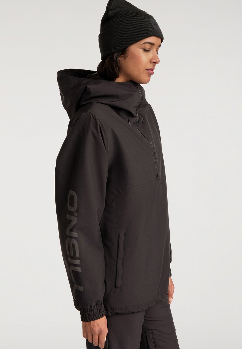 Cazadora negra con cuello alto, puños elásticos y cierre frontal de media cremallera. Presenta un logotipo prominente en la manga y un diseño minimalista.