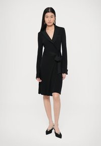 Μαύρο φόρεμα τύπου wrap με μακριά μανίκια, γιακά με εγκοπή, ζωνάκι στη μέση και λεία, ελαστική υφή. Φορεμένο με μυτερά μαύρα τακούνια.