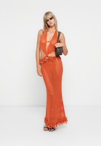 Narančasta pletena haljina s halter izrezom i otvorenim stranama, koja ima čvor na struku i resasti rub. Model drži malu crnu torbicu.