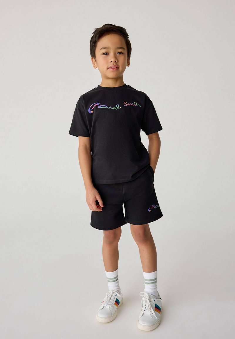 Svart bomullströja och shorts-set med flerfärgad "Paul Smith"-logga. Vita sneakers med färgglada ränder och ribbade strumpor.