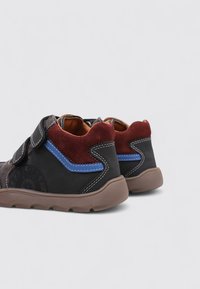 Zapatillas negras y grises con una suela marrón, detalles en gamuza roja, detalles azules y un cierre de gancho y bucle. Material texturizado visible.