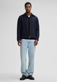 Veste zippée navy avec col, superposée à un polo noir, associée à un jean en denim bleu clair et des bottes noires.