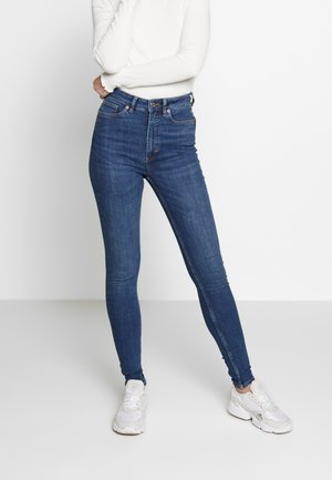 Personne portant un jean skinny taille haute bleu, un haut blanc à manches longues et des baskets blanches, debout devant un fond blanc uni.