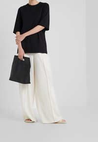 Černé oversize tričko s krátkými rukávy, spárované s širokými bílými kalhotami. Drží malou černou tašku s uchem.