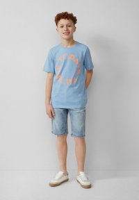 Helles blaues Baumwoll-T-Shirt mit orangem, kreisförmigem Text "DAS ZUKUNFT VON MORGEN". Getragen mit hellen Jeansshorts und weißen Sneakers mit braunen Sohlen.
