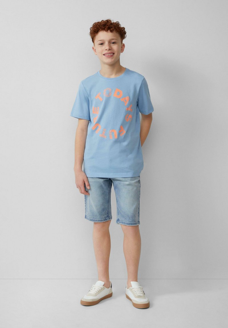 Helles blaues Baumwoll-T-Shirt mit orangem, kreisförmigem Text "DAS ZUKUNFT VON MORGEN". Getragen mit hellen Jeansshorts und weißen Sneakers mit braunen Sohlen.
