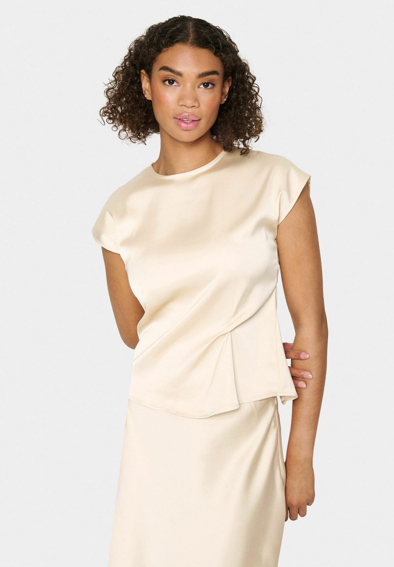 Cremfarbener Satin-Top mit kurzen Ärmeln, Rundhalsausschnitt und asymmetrischen Faltendetails an der Taille, kombiniert mit einem glatten Rock.
