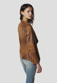 Jachetă maro din piele, cu croială lejeră, având partea frontală drapată, mâneci lungi și fante laterale. Asortată cu blugi din denim deschis la culoare.