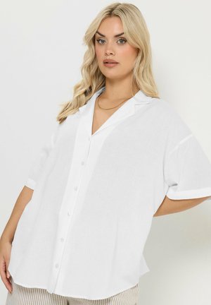 Blonde vrouw die een losse witte blouse met korte mouwen draagt, beige gestreepte broek en een dunne gouden ketting, poserend tegen een lichte achtergrond.