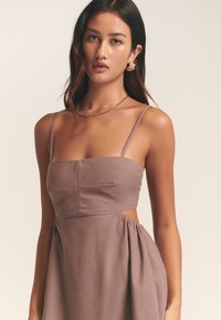 Brązowa lniana sukienka z wycięciami, cienkimi ramiączkami, dopasowanym gorsetem, marszczonym pasem i miękką fakturą; zestawiona ze złotymi kolczykami kołami.