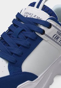 Άσπρο και μπλε αθλητικό παπούτσι Versace Jeans Couture με μπλε κορδόνια, υφασμένη μπλε γλώσσα και λογότυπο στο κέντρο και στη πλευρική επιφάνεια.
