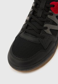 Μαύρο αθλητικό παπούτσι με πάνελ από δίχτυ και δέρμα, μαύρα κορδόνια, σόλα από καουτσούκ και κόκκινη πλεκτή λεπτομέρεια κοντά στην κορυφή.