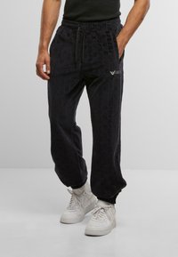 Man wearing black textured joggers with elastic cuffs and white sneakers, standing with hands in pockets against a plain background.
