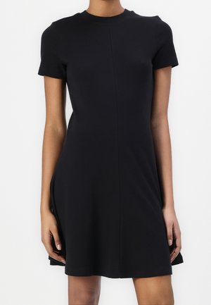 Kobieta ubrana w prostą czarną sukienkę z krótkim rękawem, okrągłym dekoltem i lekko rozszerzającym się do dołu fasonem, stojąca na białym tle.