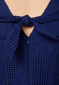 Detailní záběr na tmavě modré texturované šaty s předním zipem a uvázanou mašlí u výstřihu, která odhaluje malou oblast pleti.