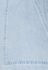 Tissu en denim bleu clair avec coutures visibles le long des coutures et des bords des poches, montrant la texture classique du jean et le motif de tissage.
