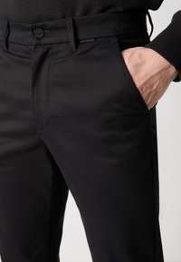 Pantalons noirs en tissu lisse, dotés d'une fermeture à bouton et de poches latérales. Le design présente des lignes épurées et une coupe ajustée.