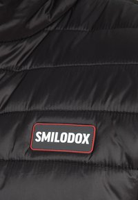 Matière de veste matelassée noire avec texture matelassée. Présente un patch logo Smilodox proéminent avec texte blanc sur fond noir bordé de rouge.