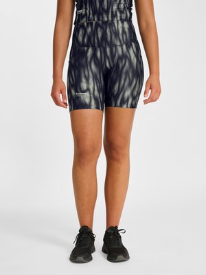 Persoon die zwarte en grijze abstracte patroon aansluitende shorts en top draagt met zwarte sportschoenen, staand tegen een effen achtergrond.