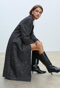 Manteau long gris foncé à motif en chevrons avec des fils blancs subtils. Doté de grands revers et d'une fermeture à boutons. Associé à des bottes noires jusqu'aux genoux.