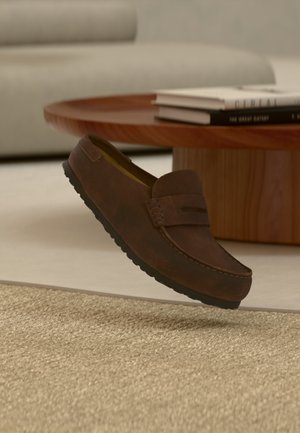 Pruun seemisnahast paeltega king, mis hõljub õhul tekstuuriga beežil vaibal, tagaplaanil ümmargune puidust laud ja diivan.