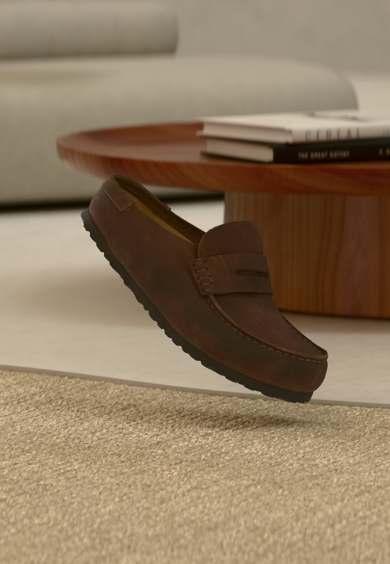 Καφέ σουέντ μοκασίνι που αιωρείται πάνω από μια υφασμάτινη μπεζ μοκέτα, με ένα στρογγυλό ξύλινο τραπέζι και καναπέ στο φόντο.