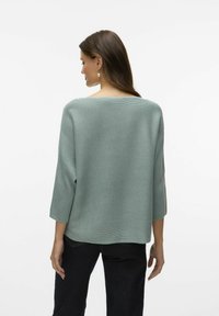 Pull en maille côtelée vert clair, avec des manches trois-quarts et un col large, associé à un jean foncé. La vue de dos montre une coupe décontractée.