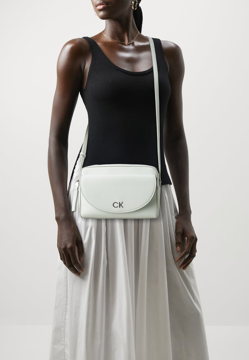 Ljusgrön läder crossbody-väska med en rundad klaff, silverfärgad hårdvara och logotyp; bärs över en svart topp, ihop med en flödande vit kjol.
