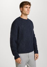 Donkerblauw gestructureerd sweater met een diamantpatroon, ronde hals en geribbelde manchetten. Gecombineerd met lichtgrijze broek, die het dikke gebreide materiaal toont.