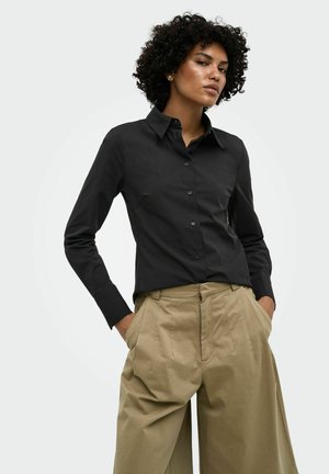 Femme aux cheveux bouclés portant une chemise noire boutonnée et un pantalon beige ample taille haute, les mains dans les poches, sur un fond uni.