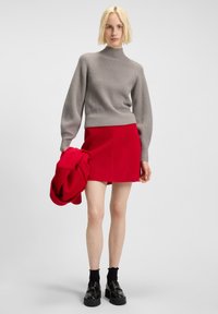 Blonde woman wearing gray ribbed sweater, red mini skirt, black socks, and platform shoes holds matching red coat in right hand.