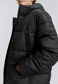 Chaqueta de plumas negra con cierre frontal, capucha con cordones y un parche de logo en la manga. Diseño acolchado texturizado.