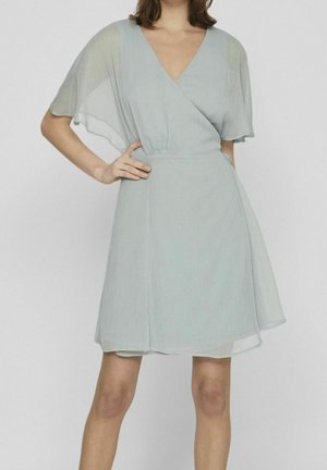 Femme portant une robe portefeuille gris clair à col en V avec des manches légères et transparentes, debout, une main sur la hanche, devant un fond uni.