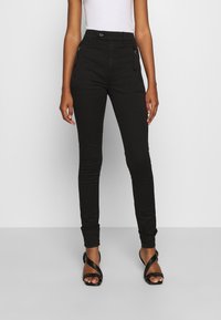 Pantalones de tiro alto negros con corte ajustado, que cuentan con bolsillos con cremallera en la parte delantera y una textura suave. Combinados con tacones negros con tiras.