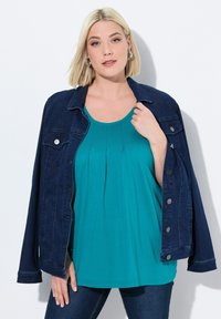 Dunkelblaue Jeansjacke mit silbernen Knöpfen, über einem tealfarbenen plissierten Oberteil getragen, kombiniert mit dunkelblauen Jeans. Glatte Texturen und lässiges Design.