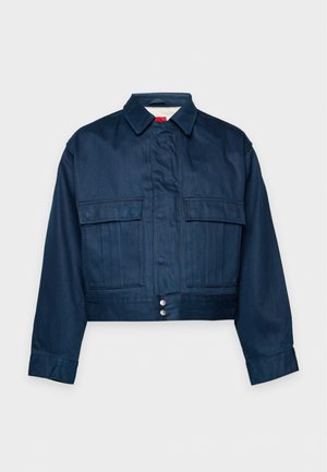 Un homme porte une veste bleu canard par-dessus une chemise jaune à carreaux, un jean foncé et des baskets beiges, sur un fond blanc uni.