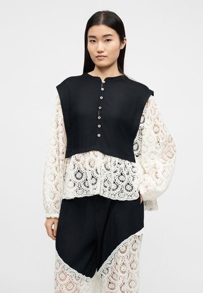 Femme portant un gilet court noir boutonné sur un chemisier blanc en dentelle à manches bouffantes, assorti à un pantalon noir avec des panneaux en dentelle.