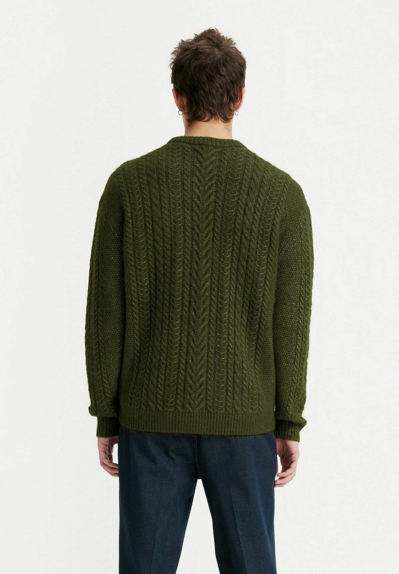 Grüner Strickpullover mit strukturiertem Kabeldesign. Verfügt über einen Rundhalsausschnitt und gerippte Bündchen. Getragen mit dunkelblauen Hosen.