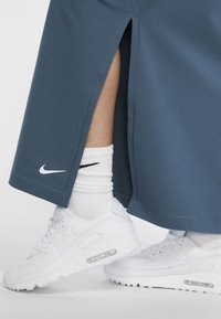 Jupe de sport bleue Nike avec fentes latérales, associée à des baskets blanches Nike Air Max et des chaussettes blanches à texture avec un détail de marque noir.