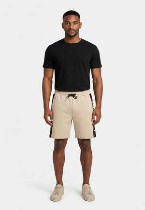 Uomo che indossa una t-shirt nera a maniche corte, pantaloncini beige con coulisse e strisce laterali nere, e sneaker beige, in piedi davanti a uno sfondo bianco semplice.