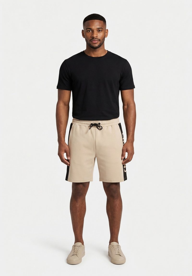 Hombre con camiseta negra de manga corta, pantalones cortos beige con cordón y franjas negras en los laterales, y zapatillas beige, de pie frente a un fondo blanco liso.
