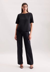 Schwarzes, kurzärmeliges Oberteil mit rundem Ausschnitt, kombiniert mit hoch taillierten, weit geschnittenen schwarzen Jeans. Das Model trägt schwarze Riemchensandalen.