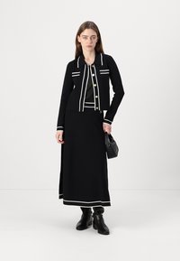 Black knit jacket with white stripes and gold buttons, paired with a black skirt featuring a white hem. Model holding a black bag.